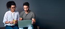 Cropped shot of two young business colleagues sitting against a gray background together and using a laptop